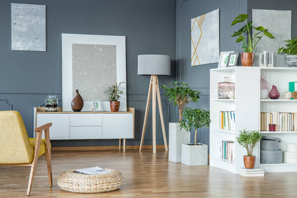 Wooden lamp placed in the corner of living room interior with fresh potted plants, gallery and books 