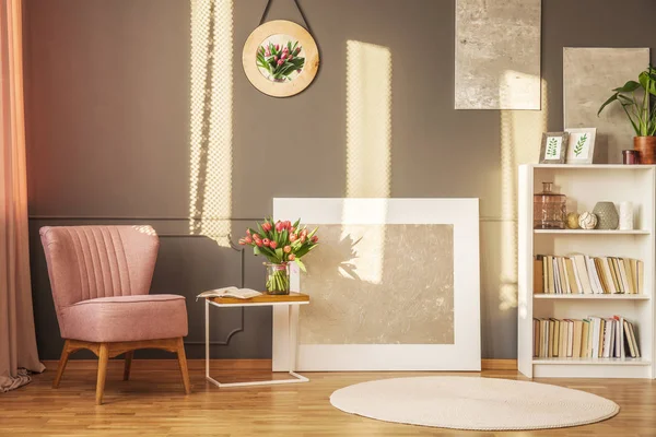 Pink armchair, flowers on a coffee table and grey painting in a sunny living room interior