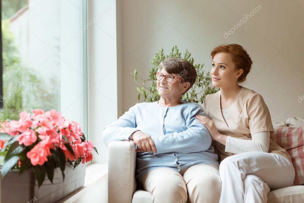 Geriatric woman and her professional caretaker sitting on a couch and looking through a window in a living room. Blooming flowers on a windowsill.