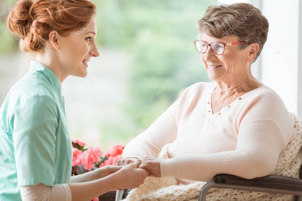 Compassionate nurse explaining a geriatric handicapped patient with dementia medical procedures while holding her hands. Assisted living house for pensioners. Side view.