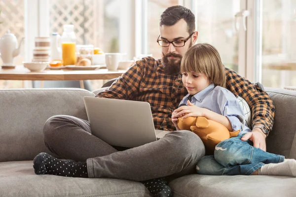 としたリビング ルームのソファの上のノート パソコンで座っているコンピューターを使用する子供を教える父 — ストック写真