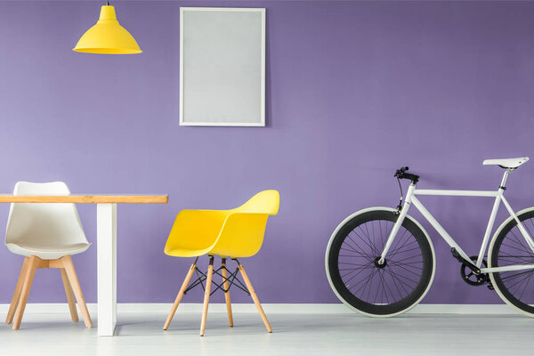 Minimal, modern interior with a white and yellow chair, a bicycle, an empty table and a hanging yellow lamp against purple background wall with a mockup poster