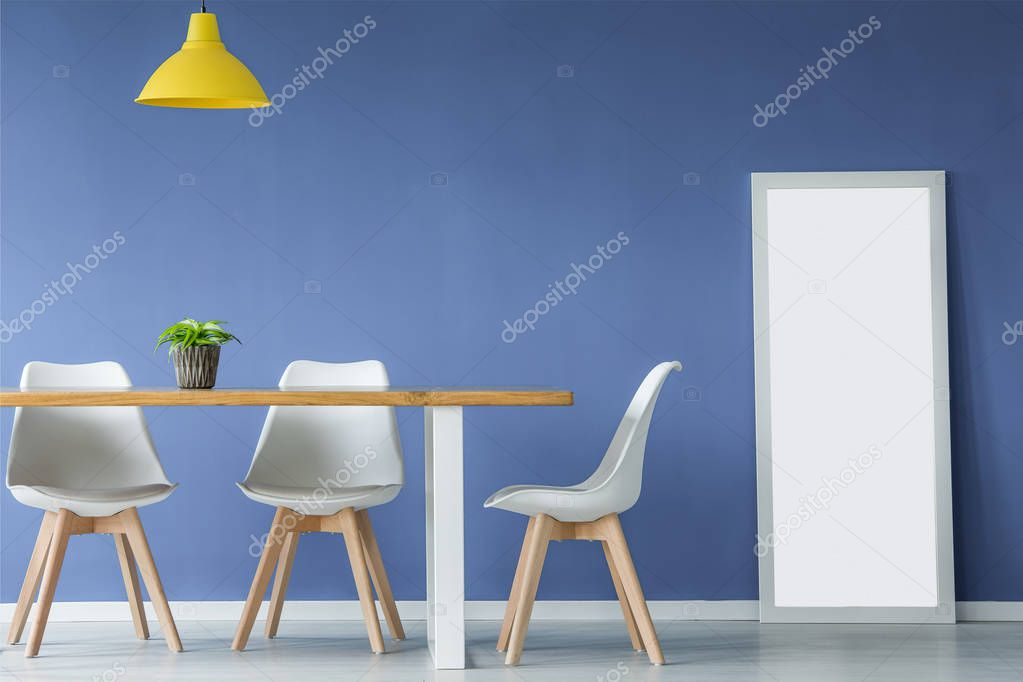 Modern open space interior with white and wooden chairs, table with a plant on top, yellow lamp and a mirror standing against blue wall