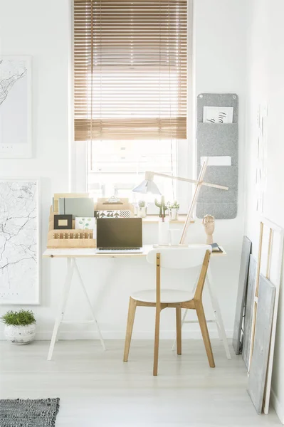 Home office desk with notebooks, lamp and laptop standing under the window with blinds in white living room interior
