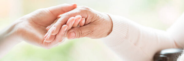 Close-up of tender gesture between two generations. Young woman holding hands with a senior lady. Blurred background. Panorama.