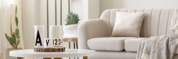 Close-up photo of white vessels standing on white end table in bright living room interior with sofa and plants in blurred background