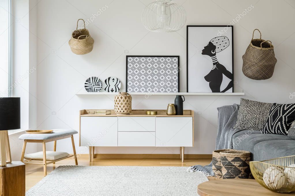 Posters and plates above wooden cupboard in boho living room interior with grey sofa. Real photo