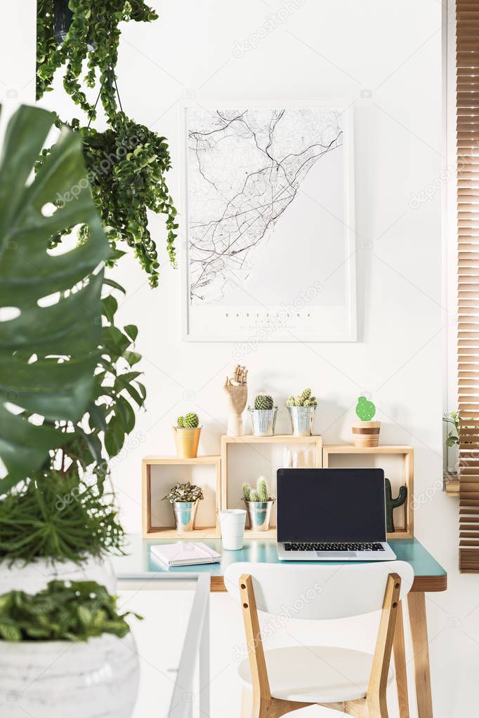 White chair standing by a simple wooden desk with cactuses, coffee cup, notebook and laptop in bright home office interior with map poster