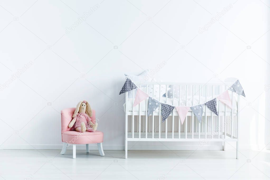 Plush toy on pink chair next to white crib in white baby's bedroom interior. Real photo