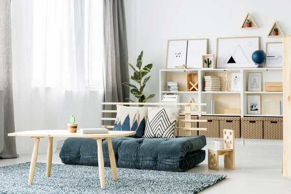 Dark green mattress sofa with two decorative cushions placed in Nordic style girl room interior with windows and wooden rack with books and posters