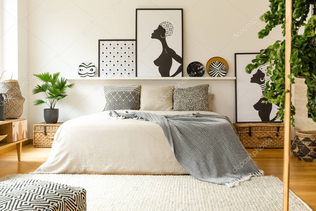 Posters and patterned plates above bed in scandi bedroom interior with plant and pouf