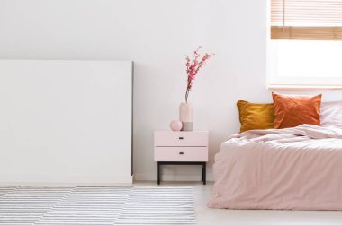 Real photo of a pink, feminine bedroom interior with orange cushions on bed and flowers on a nightstand next to an empty wall