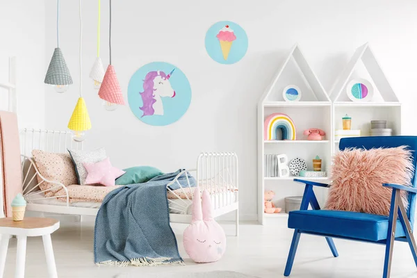毛茸茸的粉红色枕头在一个充满活力的蓝色扶手椅在一个甜美的孩子卧室内部与舒适的床上用品和卡通海报在白色的墙壁上 — 图库照片