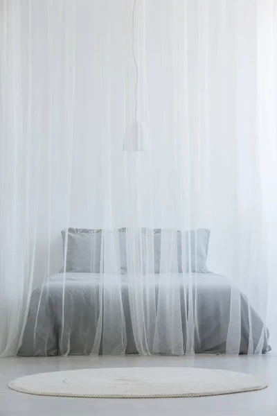 Mosquito net bed with grey bedding standing in white room interior with round rug on the floor