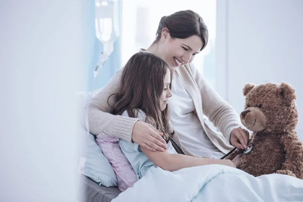 微笑的母亲和微弱的女儿照看泰迪熊在诊所 — 图库照片