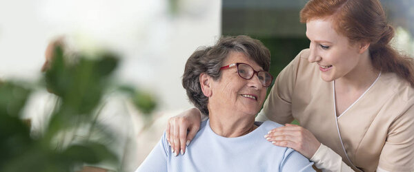 Close-up of a tender caregiver with her hands on the shoulders of a senior woman inside her home. Blurred surrounding. Panorama.