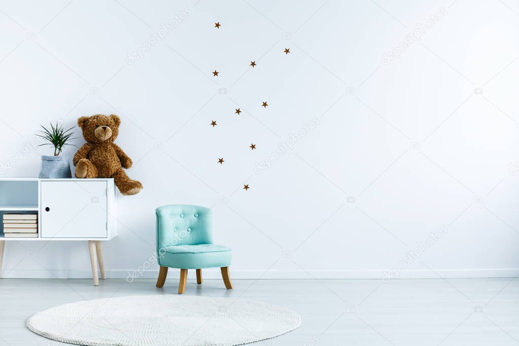 Small light blue armchair for kid standing in white room interior with stars on the wall, white rug and cupboard with books, teddy bear and fresh plant. Empty space for your crib