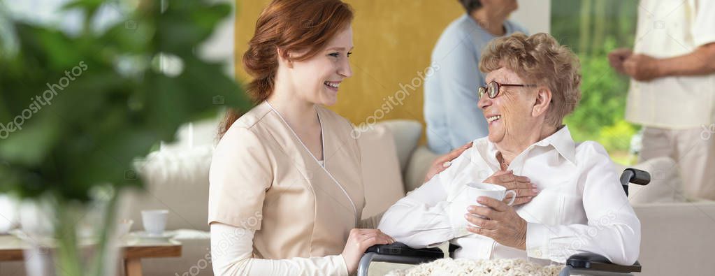 Young caretaker making a geriatric pensioner in a wheelchair laugh during leisure time in a common room of a luxury rehabilitation center. Blurred surrounding. Panorama.