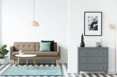 Beyaz açık alan oturma odası iç ahşap masa halı, gri dolap, basit poster ve pastel pembe lambalar ile ayakta yeşil yastıkları ile kahverengi kanepe