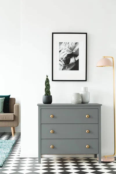 Black and white poster hanging on the wall above grey cupboard with decor standing in white room interior with checkerboard linoleum floor