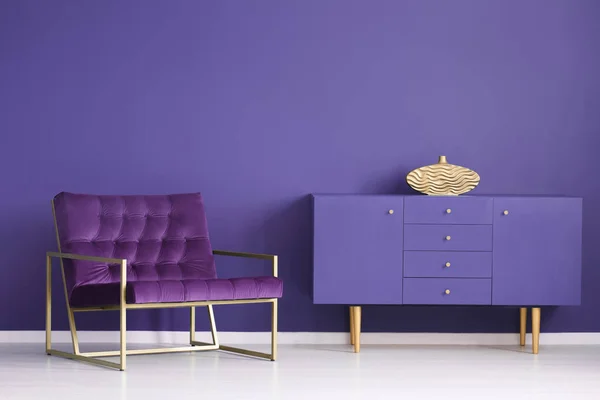 Gold vase on violet cabinet in sophisticated living room interior with purple armchair