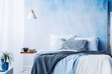 Yatak en az yatak odası iç üretim tesisi ile mavi ve gri bedsheets ile yanındaki tablosunun üst kısmındaki beyaz lamba