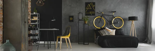 Dark studio flat interior with textured grey wall, black and yellow bike standing on bedhead, desk with geometric chair and double bed with black bedding