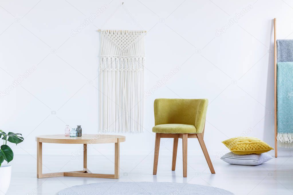 Real photo of a yellow chair standing next to a wooden table in bright, simple living room interior with ladder and cushions on the floor