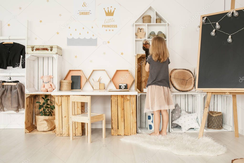 Little girl organizing her toys in white Nordic style bedroom interior with posters, wooden furniture, fluffy rug and blackboard with lights