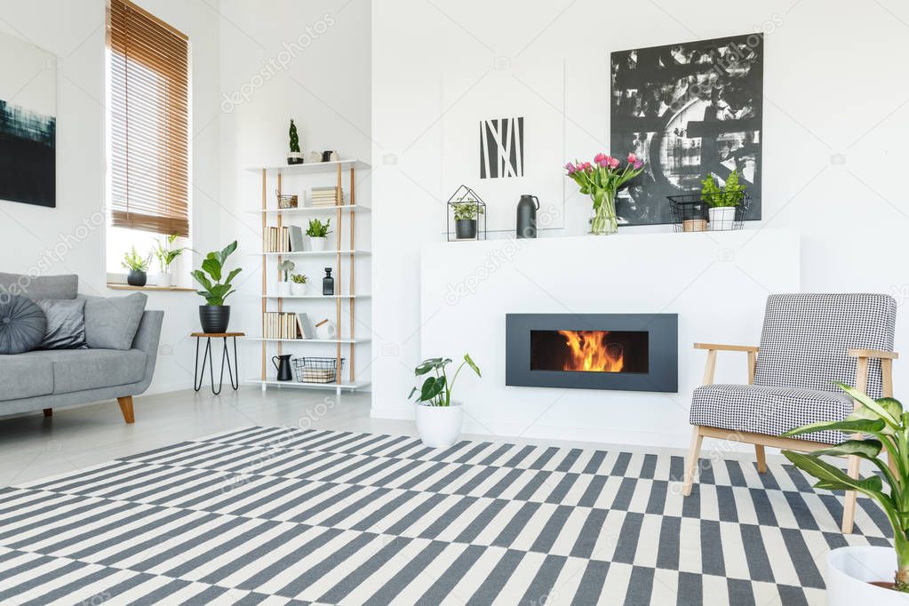 Real photo of a patterned armchair standing on a striped rug and next to a bio fireplace in warm living room interior with shelves, couch and posters