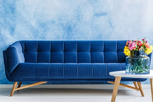 舒适的海军蓝色长毛绒沙发和五颜六色的花朵在一个花瓶上的桌子上对贝尔奥伯尔墙在客厅内部 真实照片 — 图库照片