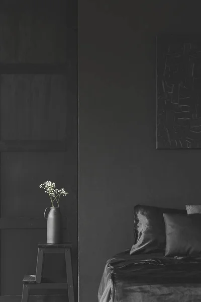 Flowers on a stool next to bed in simple black bedroom interior with dark poster