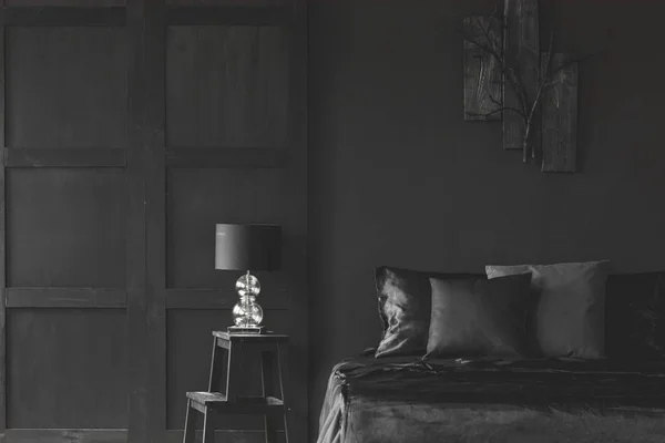 Lamp on stool next to bed in dark black bedroom interior with wooden wall. Real photo