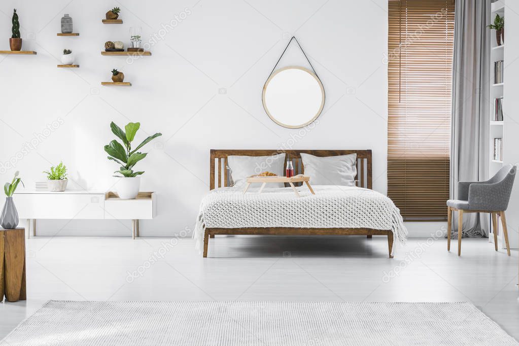 Wooden breakfast tray with croissant and juice placed on wooden double bed with white bedding standing in bright bedroom interior with fresh plants, grey chair and round mirror