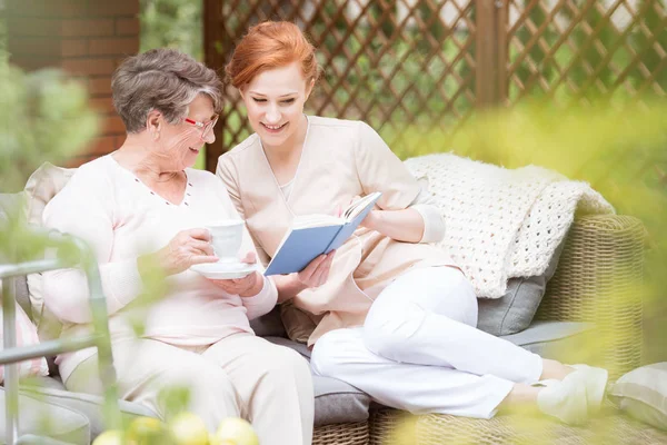 年轻的护士和一个老妇人一起读一本书在养老院的阳台上 — 图库照片