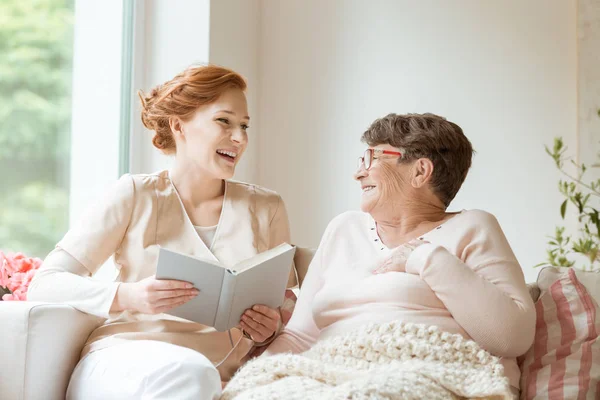 Счастливая Медсестра Читает Смешную Книгу Своему Пожилому Пациенту Частном Доме — стоковое фото