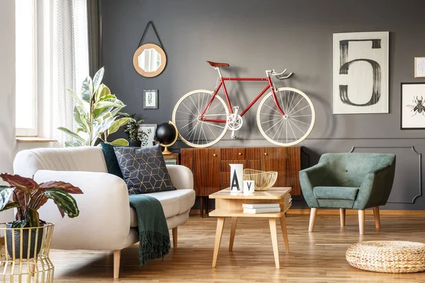 Vintage unique living room interior with white comfortable sofa with cushions and blanket, green armchair, cupboard, small table, red bicycle and a poster with number five on gray wall