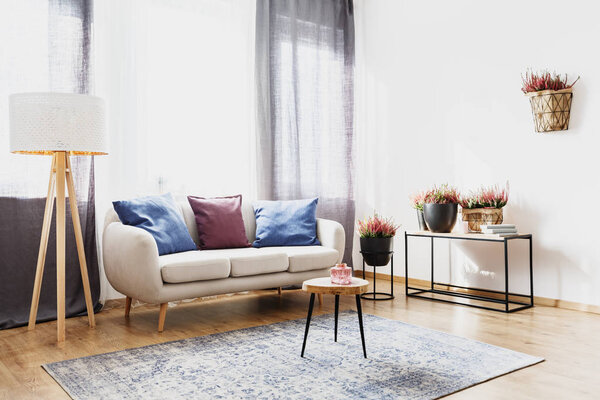 Wooden stool on carpet near lamp and sofa with purple and blue pillow in living room interior with heathers