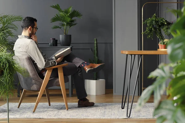 现代男子看书时坐在灰色扶手椅在时髦的室内与植物 — 图库照片