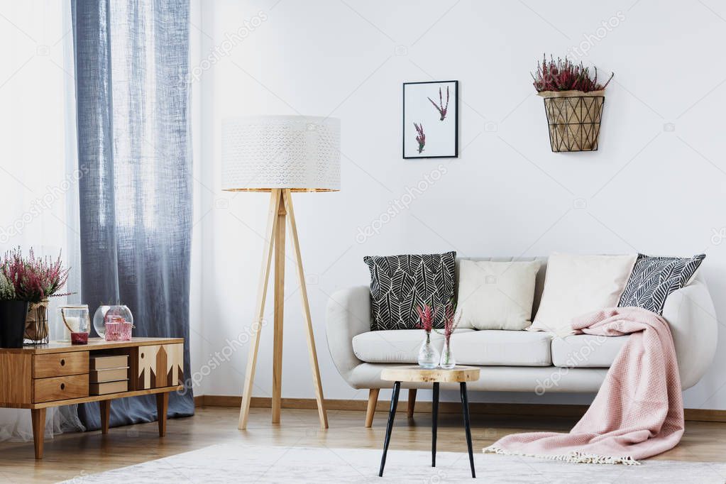Basket with heather and poster on white wall in simple living room interior with lamp, sofa, cupboard and table