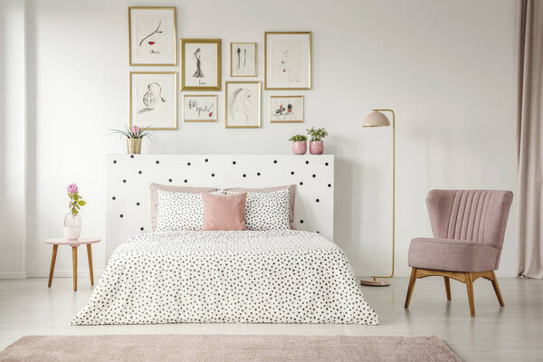 Framed posters gallery above a cozy double bed in a feminine white room interior with a comfy, retro armchair