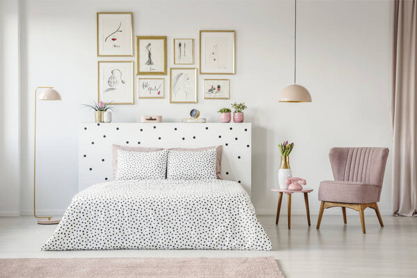 Pink armchair next to patterned bed in feminine bedroom interior with gallery of posters