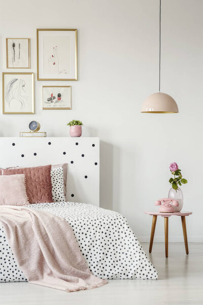 Pink, old-fashioned phone on a wooden side table and a pendant lamp in a serene bedroom interior with white walls
