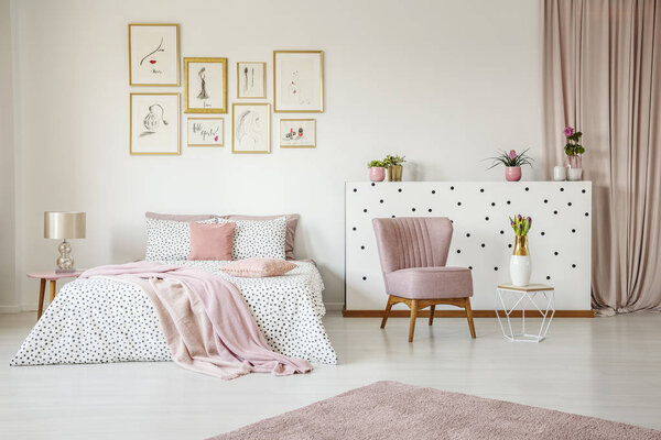 Pink armchair in spacious bedroom interior with blanket on bed against white wall with posters