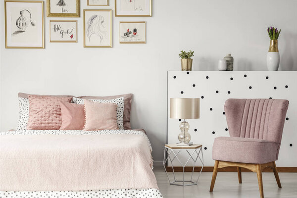 Pink armchair next to table with lamp and bed in pastel bedroom interior with posters