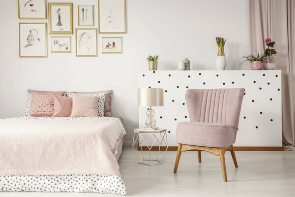 Lamp on table between bed and pink armchair in elegant bedroom interior with posters