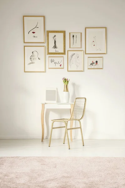 Golden chair by an elegant vanity table with a mirror by a white wall with drawings gallery in a feminine bedroom interior with a pink rug