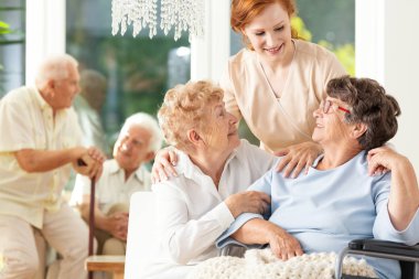 Mutlu dostluk yaşta. İhale caregiver arkasında, üst düzey kadın birbirlerini huzurevinde sarılma. Arka plan bulanık erkeklerde.