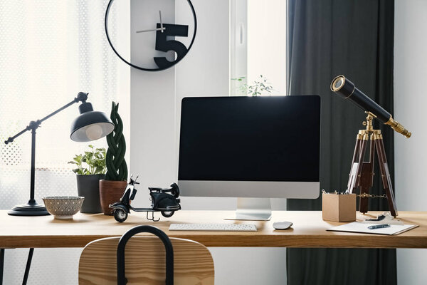 Computer with mockup placed on wooden desk with scooter model, lamp, vintage telescope and fresh plants in bright home office interior with window