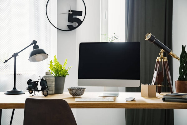 Close-up photo of computer with mockup standing on a wooden desk with vintage telescope, fresh green plants and metal lamp